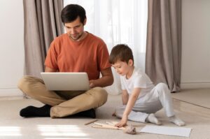 A parent and child sit on a soft carpet, engaged with a laptop, surrounded by art supplies and papers in a cozy living room.