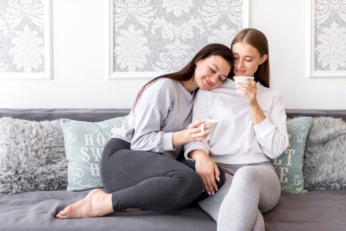 Two women sit close together on a gray couch, enjoying warm beverages, surrounded by cozy pillows and a stylish decor backdrop.