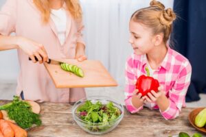 A person slices cucumbers on a wooden board, while a child holds a red bell pepper next to a bowl of mixed salad greens.