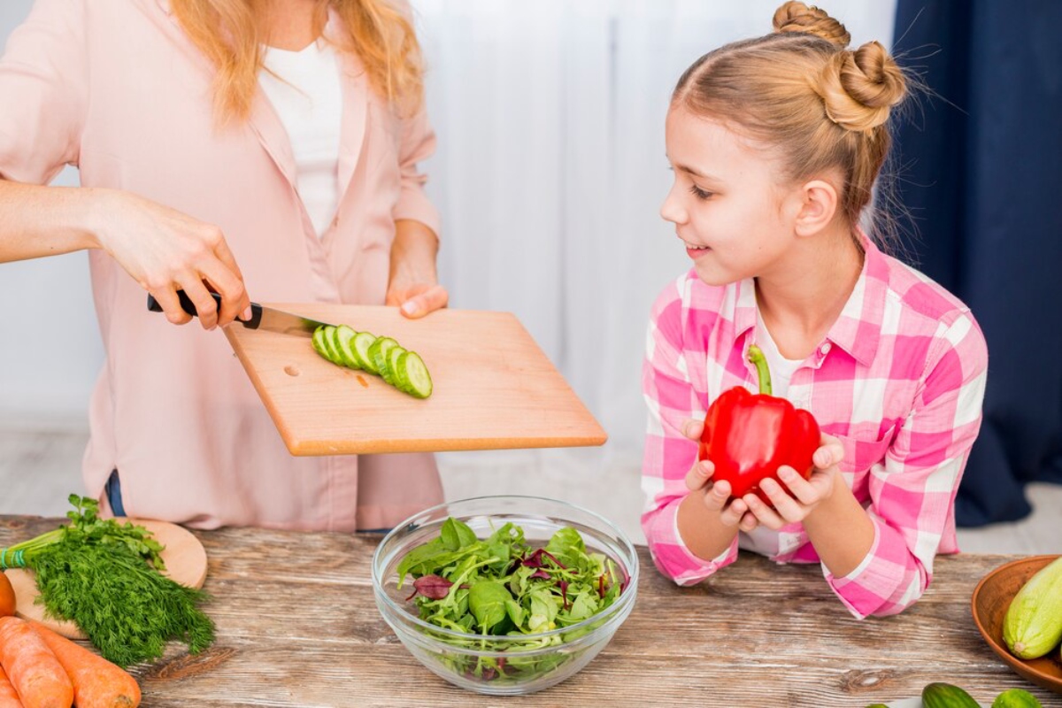 A person slices cucumbers on a wooden board, while a child holds a red bell pepper next to a bowl of mixed salad greens.