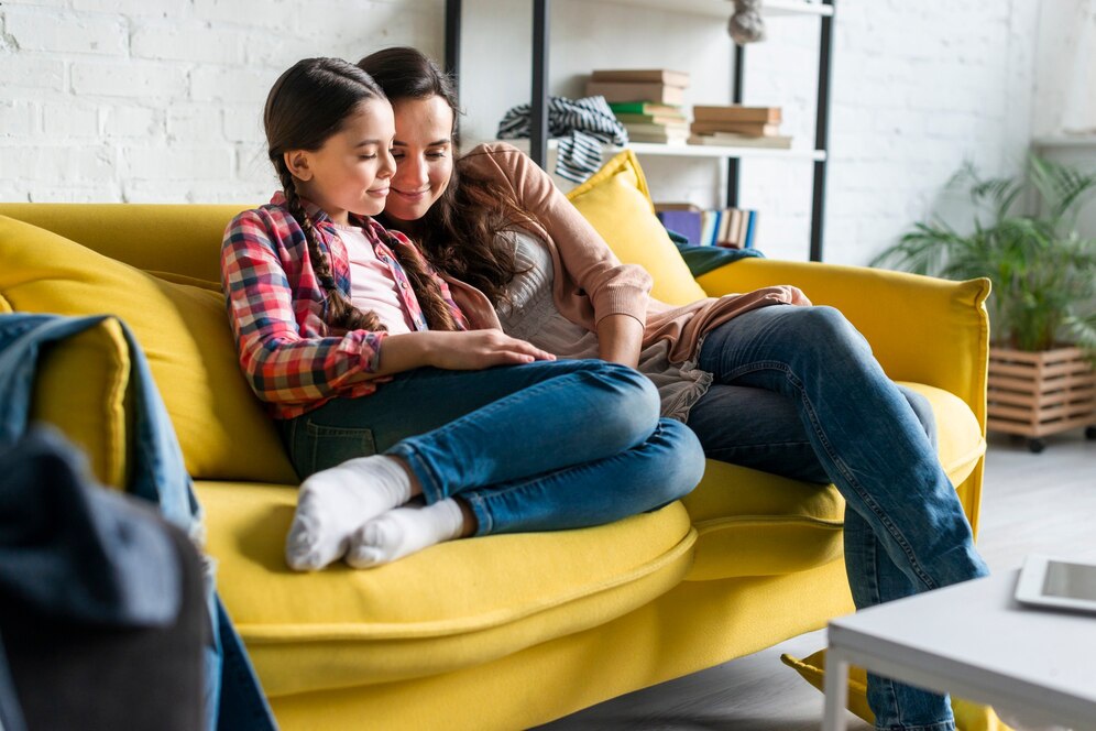 A woman and a child sit together on a bright yellow couch, sharing a moment of joy in a cozy, well-lit living room.