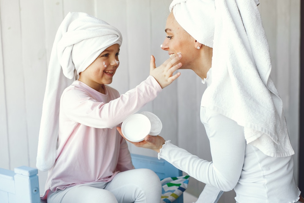 A woman and a child, both in towels, apply skincare products in a cozy bathroom setting, sharing a joyful skincare moment together.