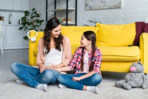 A woman and a girl sit on a soft rug, holding hands, with a bright yellow couch and a stuffed animal nearby, in a cozy living room.