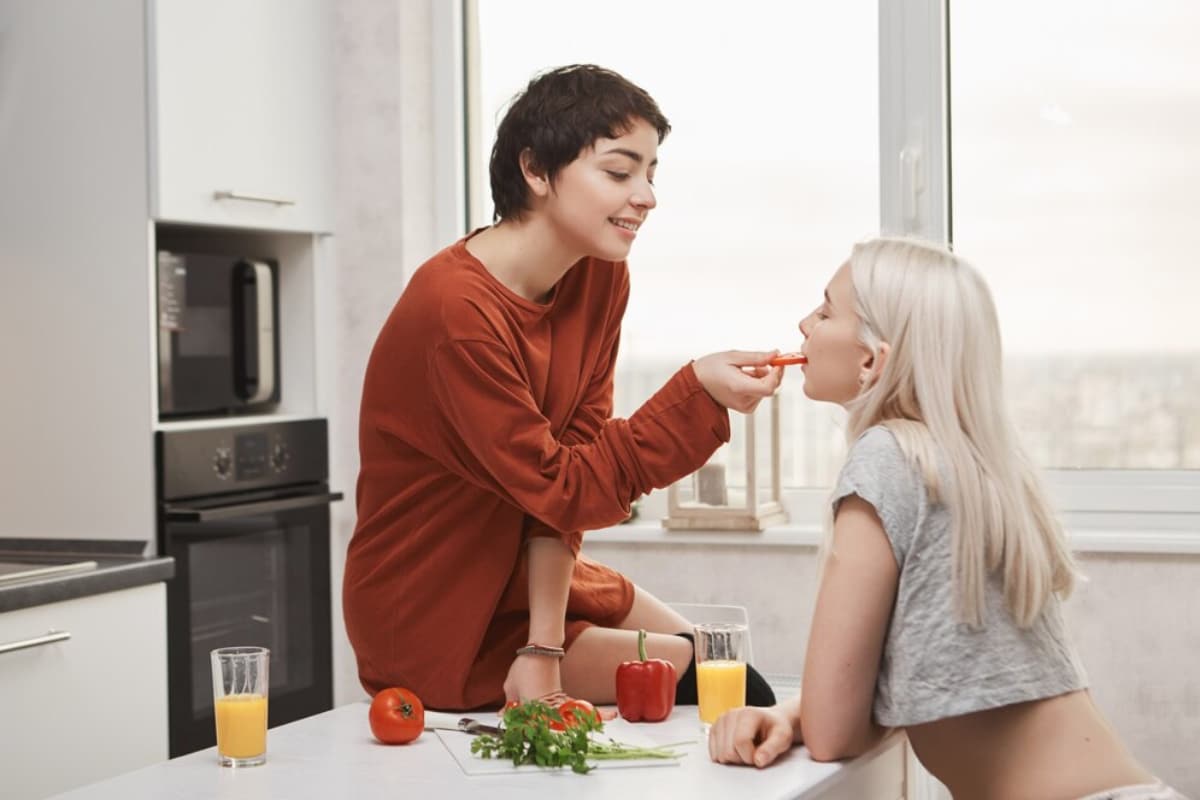 Two individuals share a moment in a modern kitchen, feeding each other fresh vegetables and surrounded by juice and ingredients.
