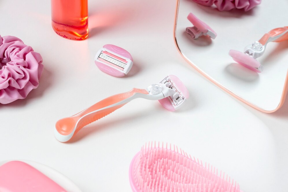 A pink-themed beauty set featuring a razor, hair scrunchie, brush, and serum bottle arranged on a clean surface.