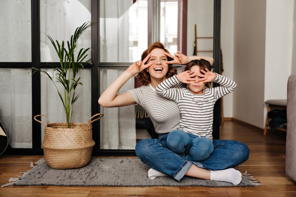 A woman and a child sit on a rug, playfully posing with peace signs, surrounded by a plant and modern home decor.