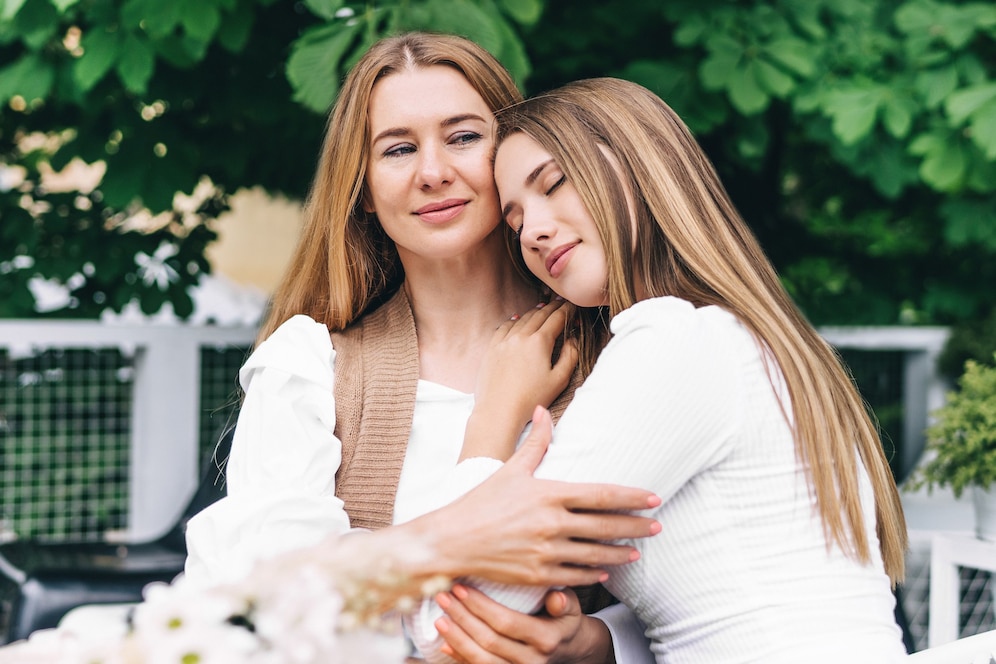 Two women share an intimate moment outdoors, surrounded by greenery, one woman gently touches the other's arm in a warm embrace.