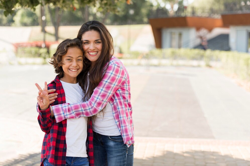 A woman and a child pose together outdoors, smiling, with the child making a peace sign. They wear colorful plaid shirts.