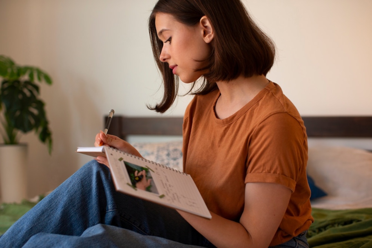 A person wearing an orange shirt sits on a bed, writing notes in a notepad, surrounded by a cozy room with a green plant.