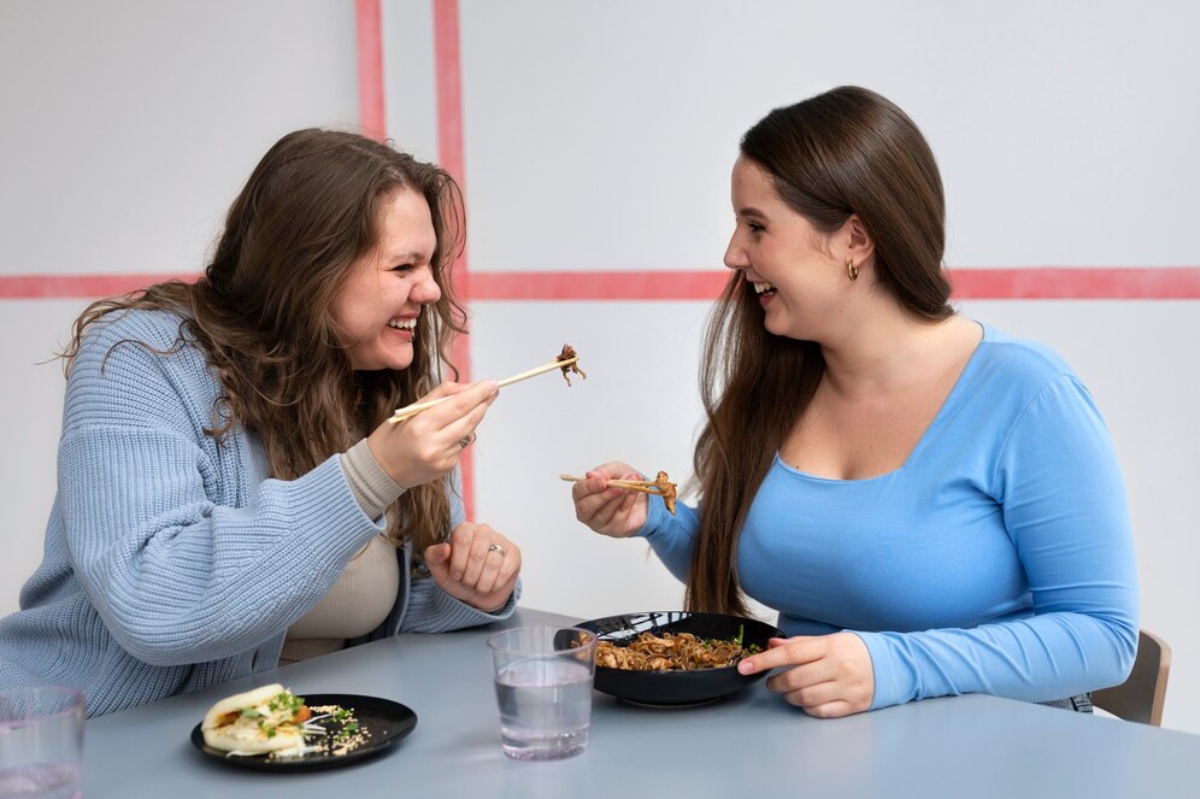 Two women share a meal, using chopsticks, at a table with a bowl of noodles and a glass of water, in a cozy, minimalist setting.