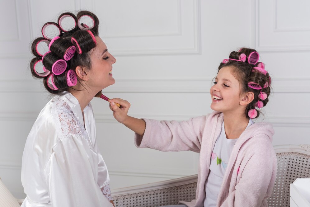 A woman in a white robe sits while a girl wearing a pink robe applies makeup, both sporting various hair curlers.