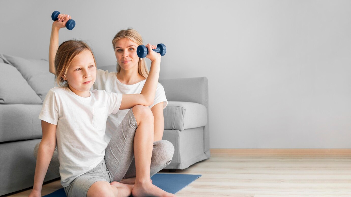 Two individuals workout at home, lifting small blue dumbbells while seated on a yoga mat, with a light gray sofa in the background.