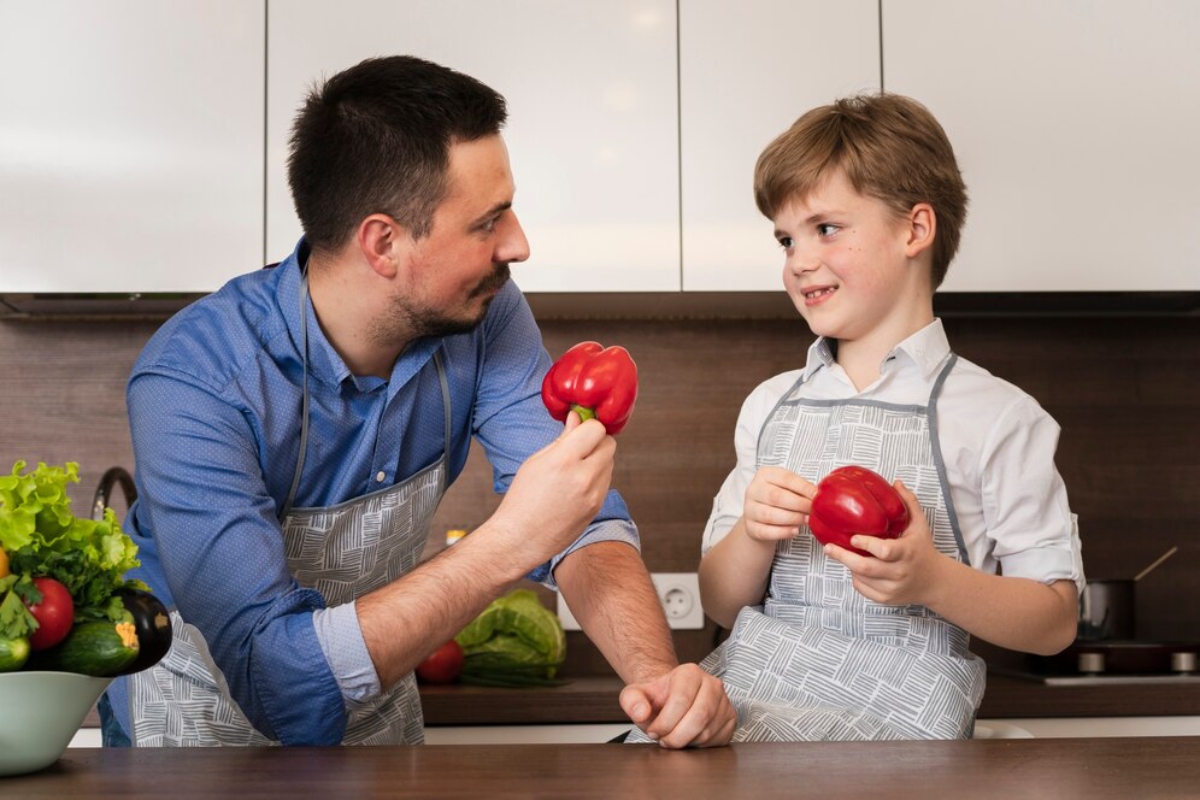A man and a boy in matching aprons hold red bell peppers while preparing ingredients in a modern kitchen filled with vegetables.