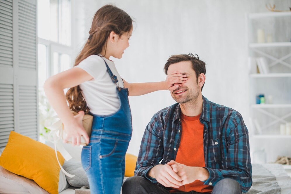 A child in a white shirt and blue overalls stands beside a man, both engaged in conversation in a bright living room.