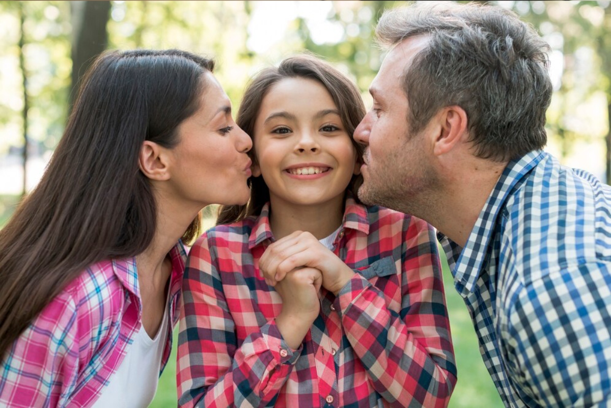 Two adults lean in close to a young girl in a park, all wearing plaid shirts, with a warm and affectionate atmosphere.