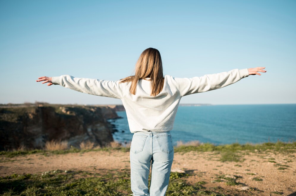 A woman stands with arms outstretched on a cliff, overlooking a serene blue ocean under a clear sky.