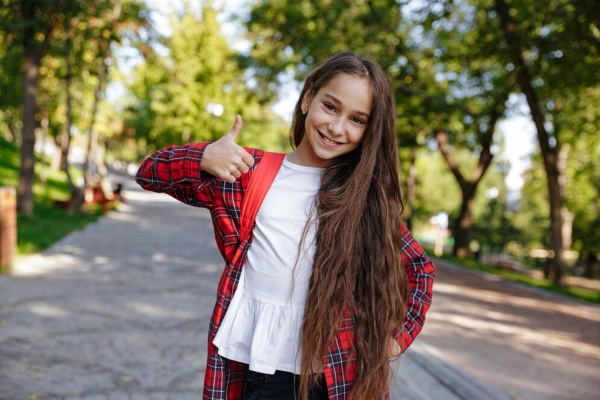 A young girl wearing a red and black plaid shirt stands outdoors, giving a thumbs-up gesture with a cheerful expression.