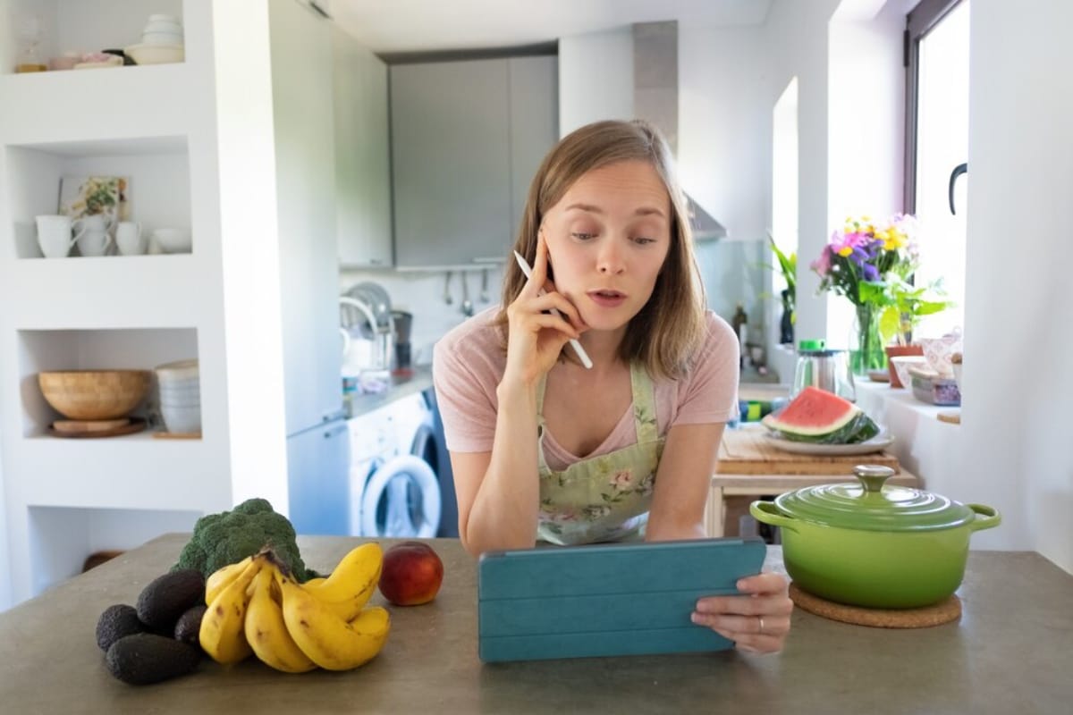 A woman in an apron, standing in a bright kitchen, reviews a tablet with fresh fruits and vegetables on the counter.
