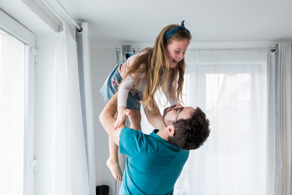 A man joyfully lifts a young girl in a bright, airy room with large windows and light curtains, showcasing a playful bonding moment.