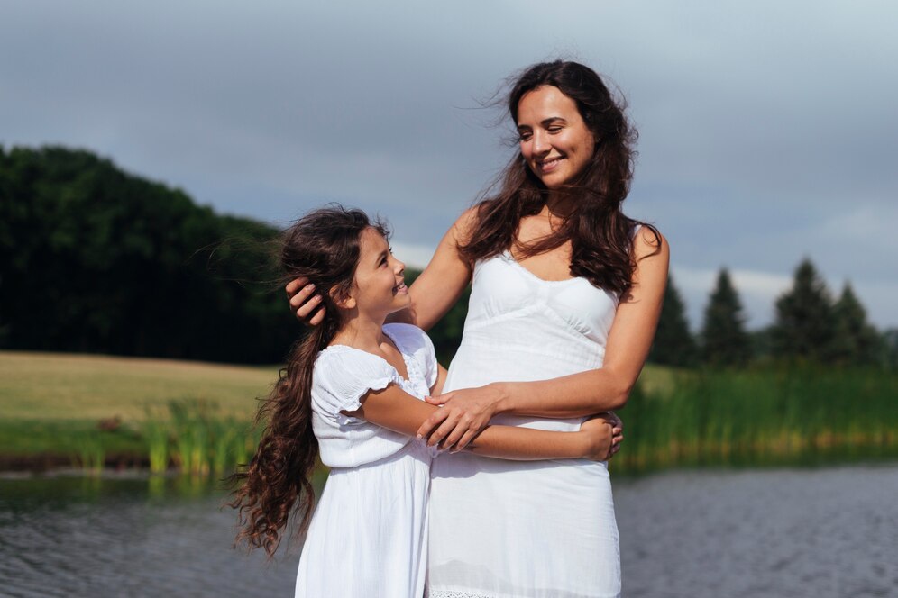 A woman and a girl embrace by a serene pond, both wearing white dresses, with lush greenery and a cloudy sky in the background.