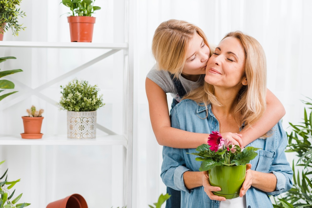 A woman holds a potted flower while a girl embraces her from behind, surrounded by various indoor plants on shelves.