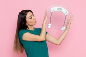 A person in a green t-shirt holds a modern, clear bathroom scale against a pink background, smiling confidently.
