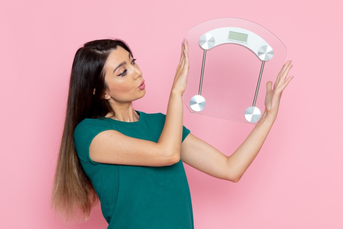 A person in a green t-shirt holds a modern, clear bathroom scale against a pink background, smiling confidently.
