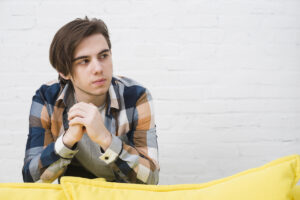 A young man in a plaid shirt sits pensively on a yellow couch against a white brick wall, hands clasped together.