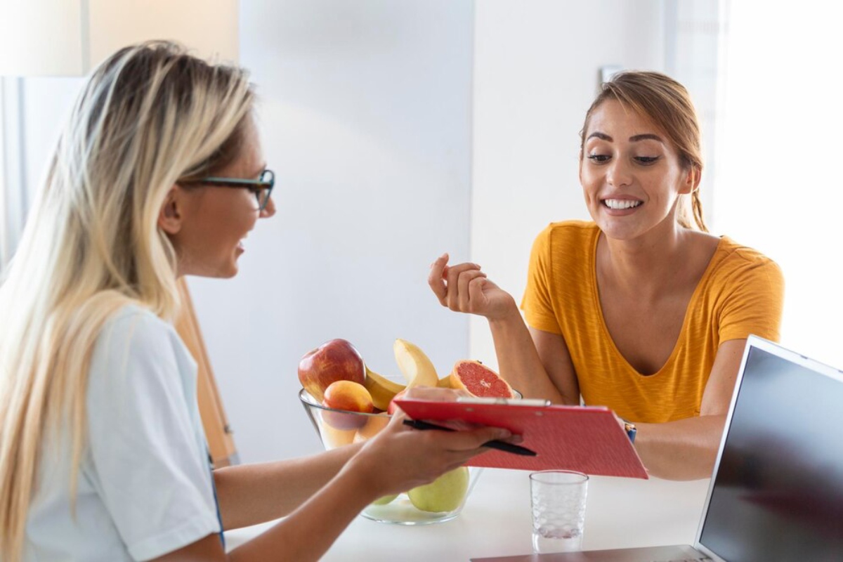 A woman in a white shirt discusses health with another woman in a yellow top, surrounded by fresh fruit and a laptop.
