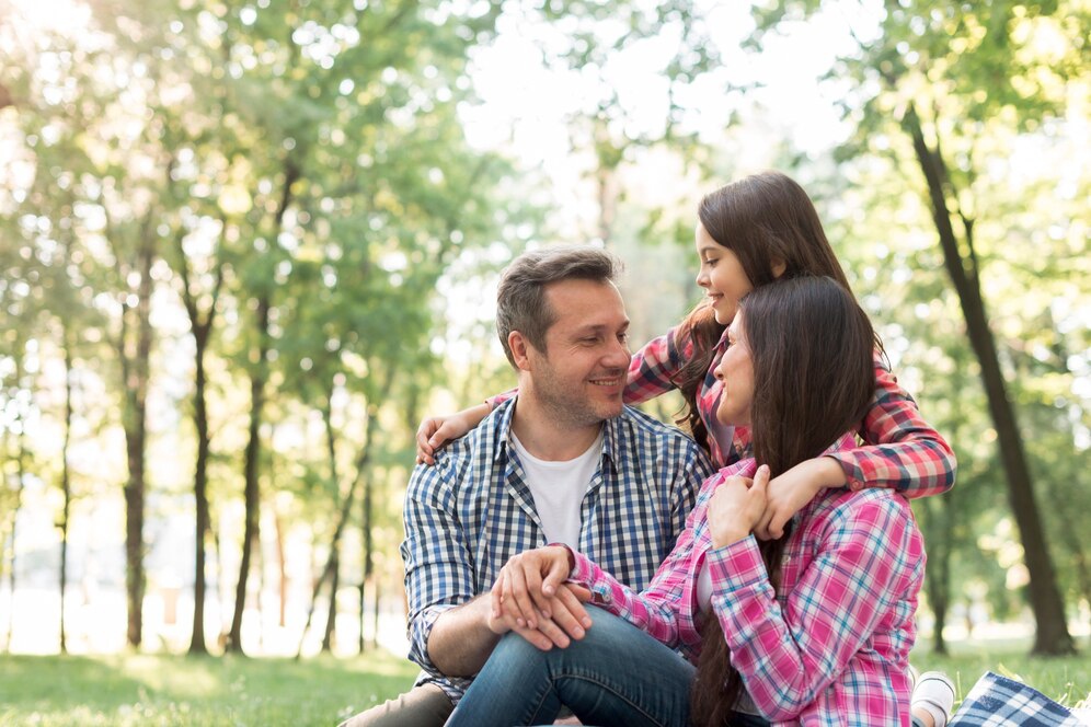 A man and two individuals sit together on grass in a sunlit park, surrounded by lush trees, sharing a joyful moment.