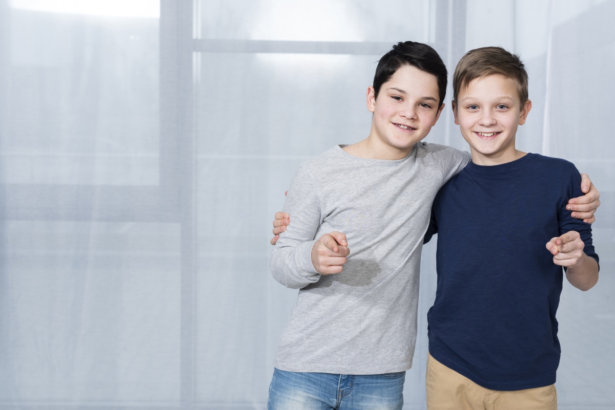 Two boys stand close together, smiling and pointing at the camera, in a bright room with sheer curtains.