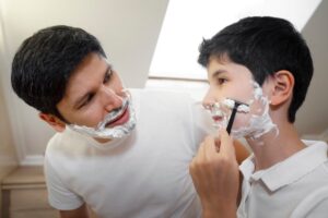 Two individuals in white shirts are engaged in shaving, with shaving cream on their cheeks and a well-lit, indoor setting.