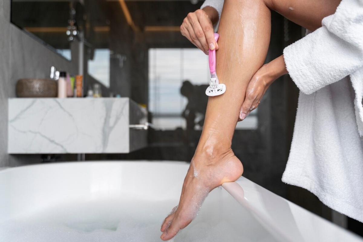 A woman in a white bathrobe shaves her leg with a pink razor in a modern bathroom with a tiled backdrop and plush soaking tub.