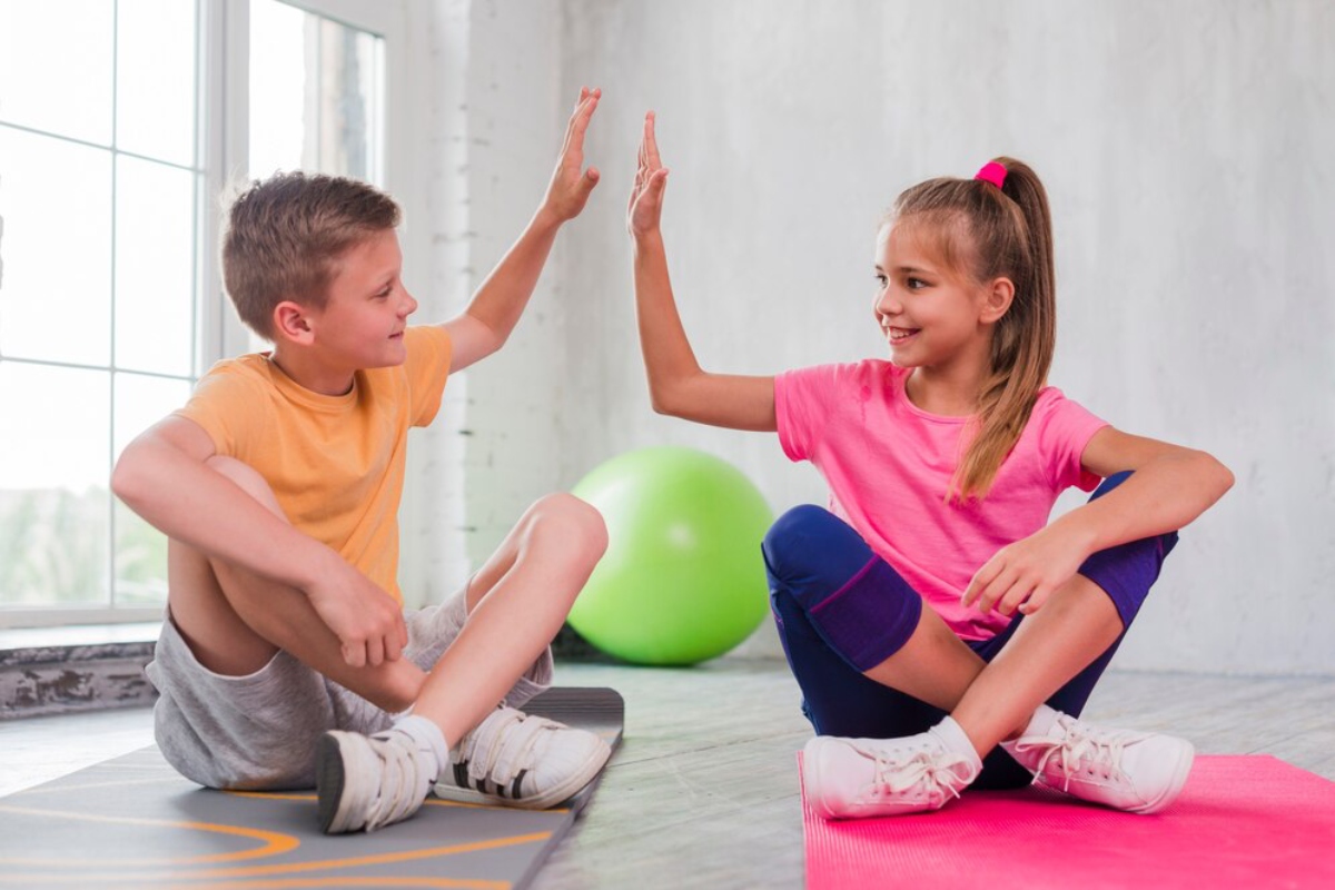 Two children, one in an orange shirt and the other in pink, sit on yoga mats, hand-slapping in a playful interaction.
