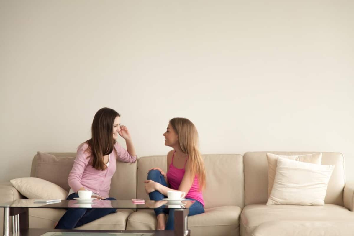 Two women sitting on a light-colored sofa, engaged in conversation, with coffee cups and a minimalistic decor around them.