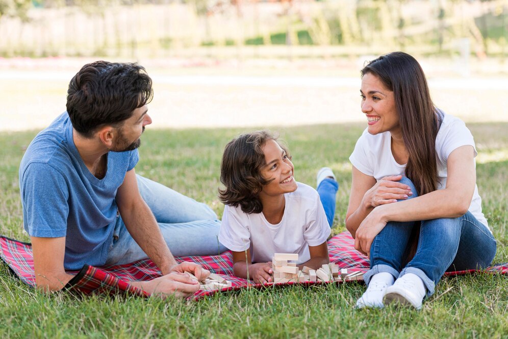 A family enjoys a leisurely afternoon on a picnic blanket in a grassy park, playing with wooden blocks together.