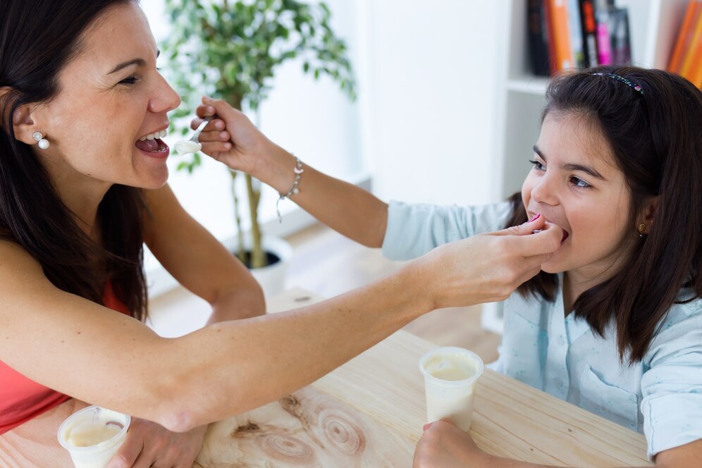 A woman feeds a child a spoonful of dessert at a wooden table, surrounded by green plants and bright, modern decor.