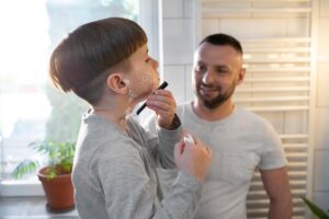 A child in a gray shirt mimics shaving in a bright bathroom, holding a marker and cream, with a plant nearby.