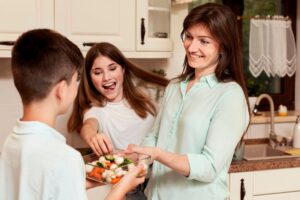 A person hands a glass dish of colorful vegetables to a child in a bright kitchen, with another child reaching for the dish.