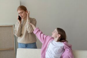Two women are interacting in a cozy indoor setting; one is speaking on the phone while the other reaches out, wearing a pink cardigan.