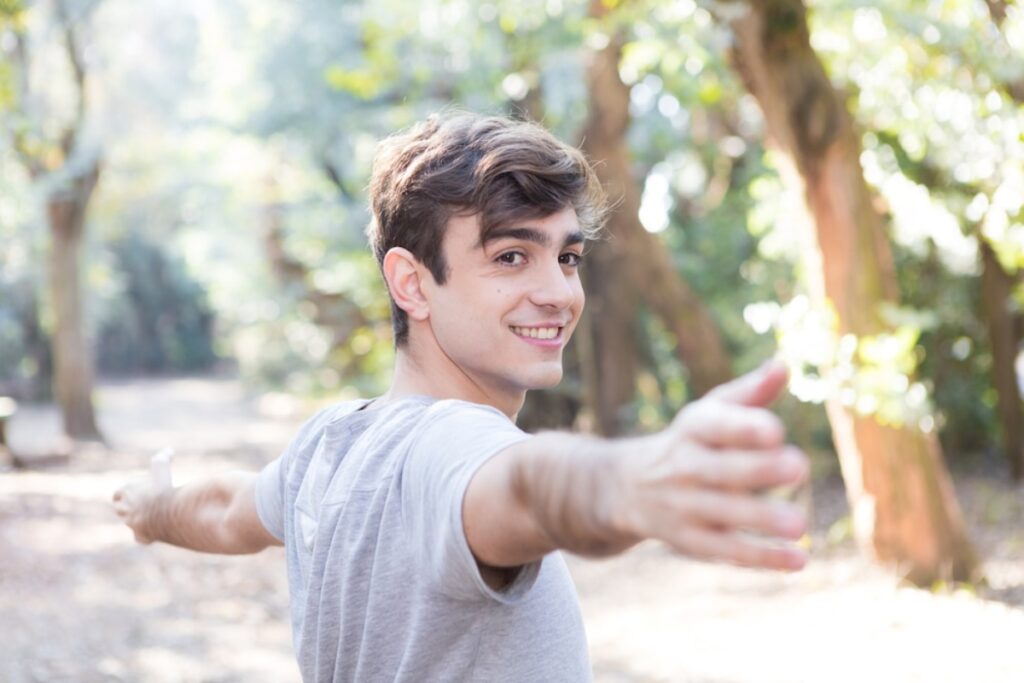 A person with arms outstretched stands in a sunlit forest, surrounded by lush greenery and soft, dappled sunlight.