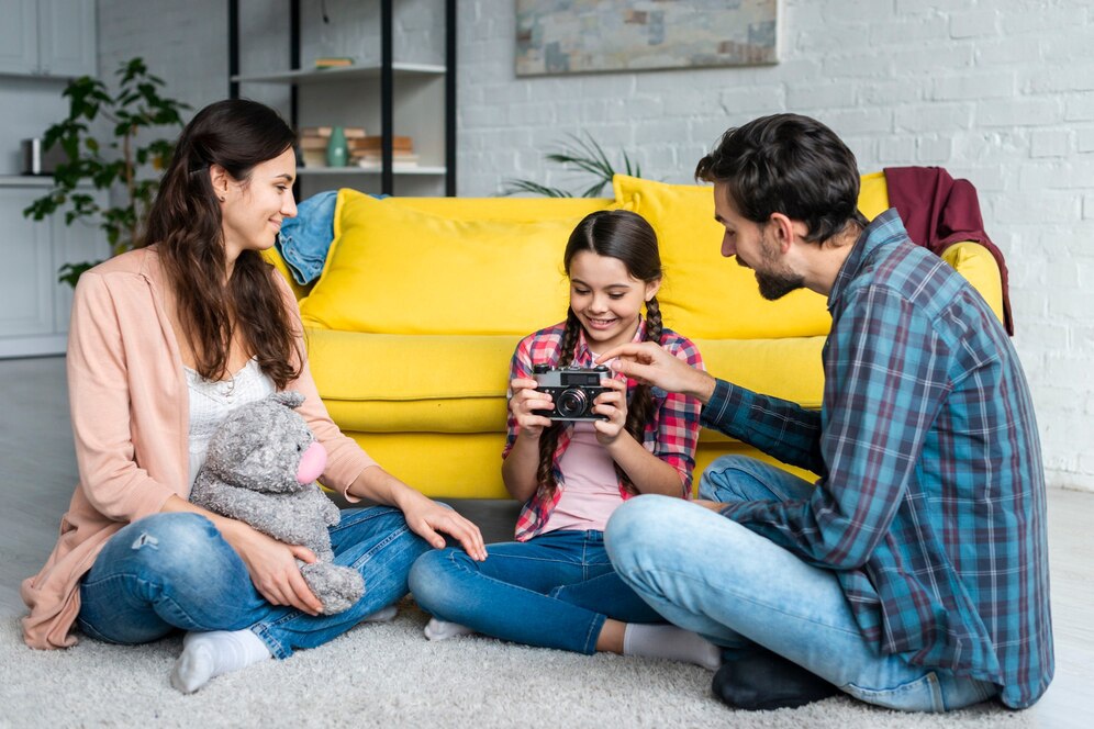 A woman and a man engage with a girl sitting on the floor, as she holds a camera, with a yellow sofa in the background.