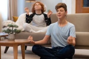 Two children practice meditation in a cozy living room, sitting cross-legged and calmly focused, with a small potted plant nearby.