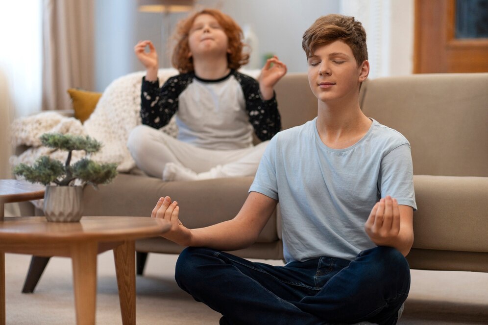 Two children practice meditation in a cozy living room, sitting cross-legged and calmly focused, with a small potted plant nearby.