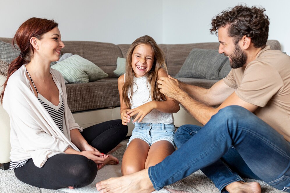 A family sits on a rug in a cozy living room, engaging in a playful interaction with a young girl in casual clothing.