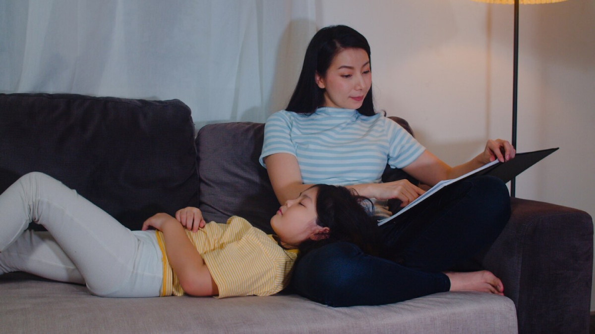 A woman in a striped shirt sits on a couch, reading a book with a child lying beside her, both enjoying cozy family time.