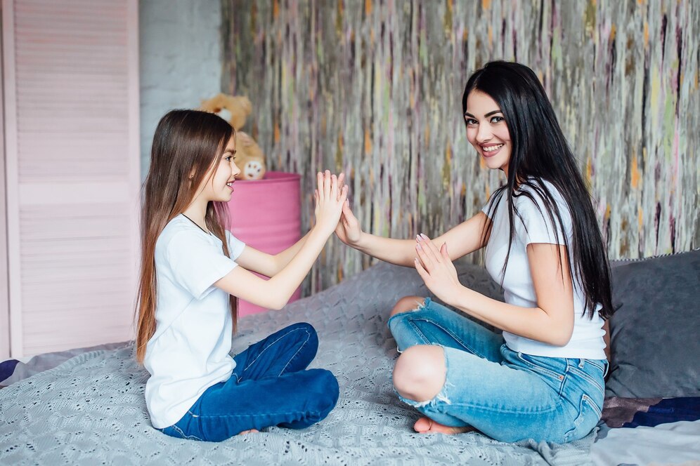 A woman and a girl sit on a bed, playing a hand-clapping game, surrounded by a cozy room with a colorful patterned wall.