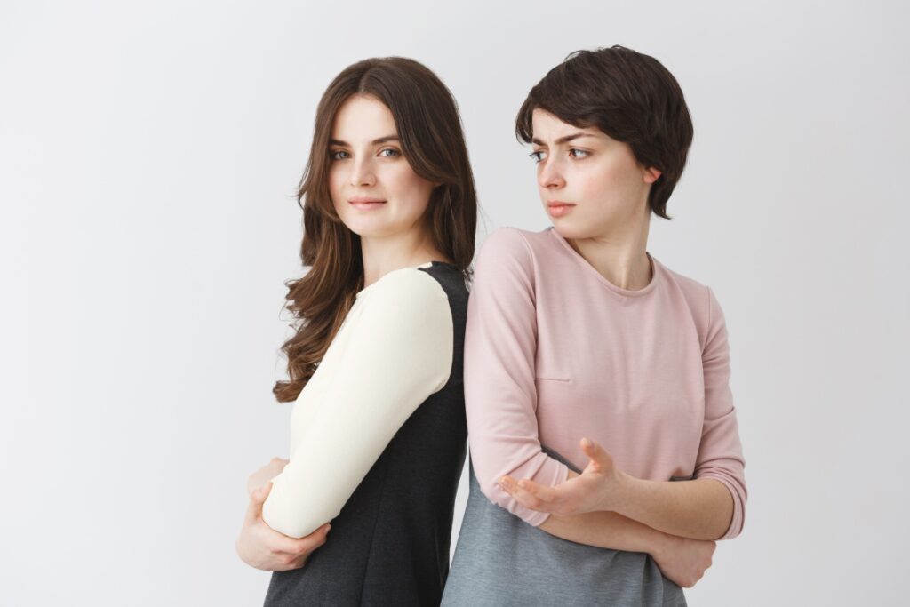 Two women standing back-to-back with crossed arms; one is smiling confidently while the other looks skeptical or annoyed.
