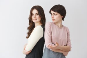 Two women standing back-to-back with crossed arms; one is smiling confidently while the other looks skeptical or annoyed.