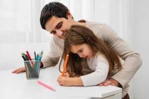 A child with long hair writes on paper at a table, while an adult gently supports them. A container of colored pencils is nearby.